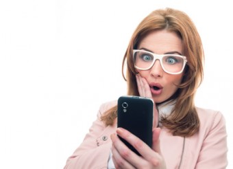 Amazed young woman looking her mobile phone, isolated on white. Trendy girl using smartphone, studio shot