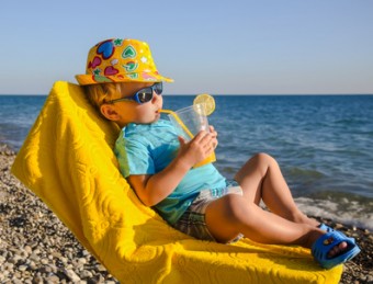 Boy kid in armchair with juice glass on beach against sea