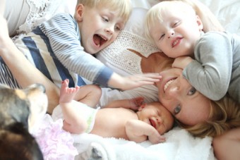 A happy young mother is laying on the bed with her three young children, two wild boys and a newborn baby girl, as their pet dog sits by.