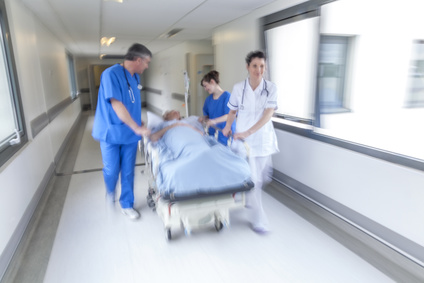 A motion blurred photograph of a senior female patient on stretcher or gurney being pushed at speed through a hospital corridor by doctors & nurses to an emergency room